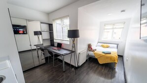 A compact hotel room with a bed, a desk with a laptop, a chair, a window with blinds, and a small table. - Stunning studio next to restaurants! (Birmingham)