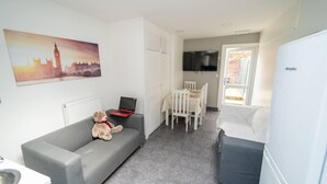 A compact living space with a grey sofa, a teddy bear, a laptop, a dining table with chairs, and a TV mounted on the wall. - Stunning studio next to restaurants! (Birmingham)