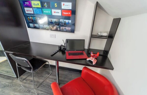 A modern office space with a desk, laptop, and gaming controller. A TV mounted on the wall displays various streaming services.