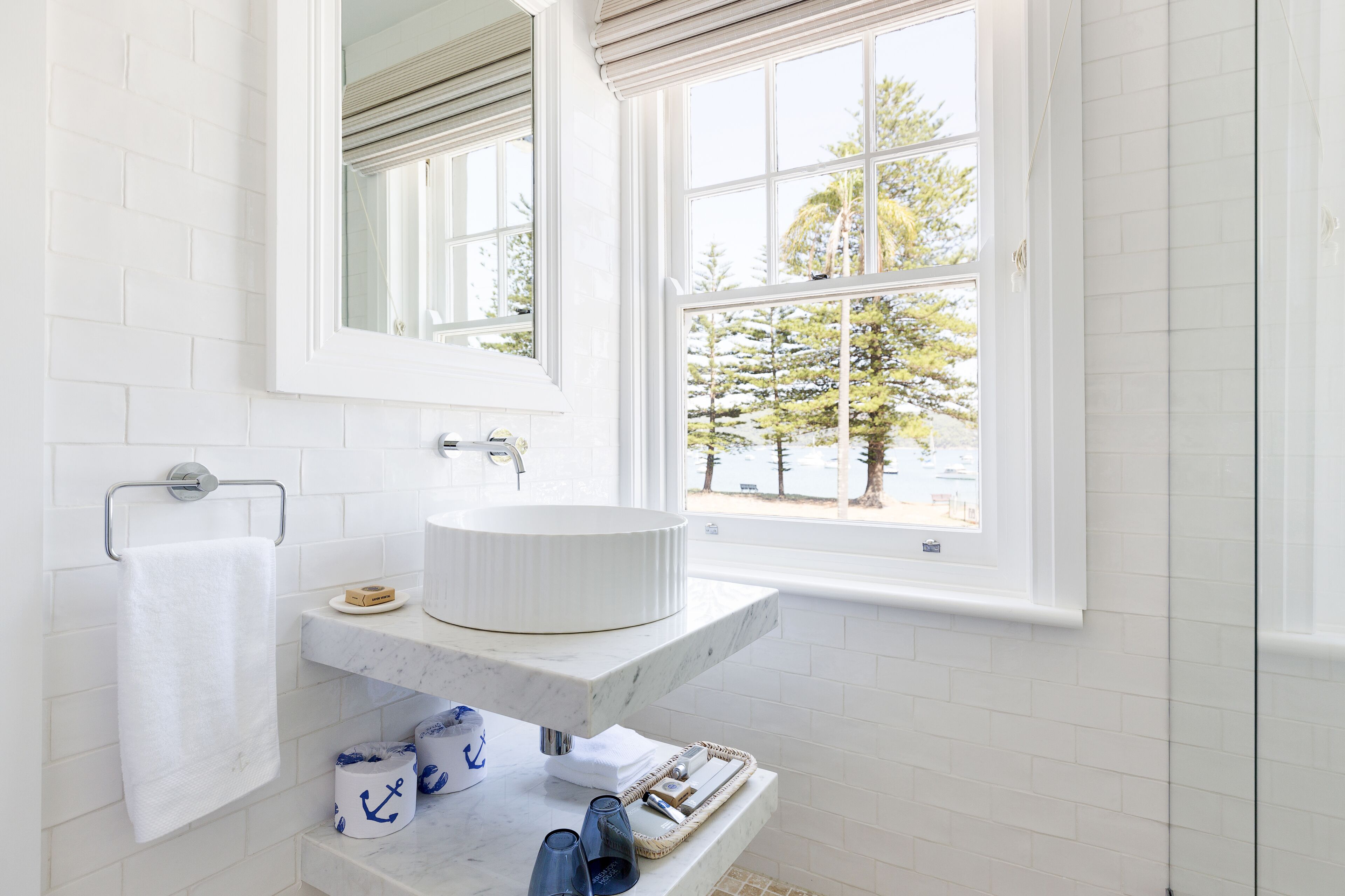 Une salle de bain avec un lavabo blanc, un porte-serviettes et une fenêtre donnant sur des arbres et un immeuble.