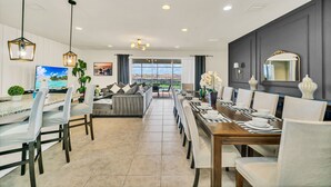 A modern dining area with a long table set for six, a sofa, and a TV on the left. - 9Br/6Ba lake view Pool House from 330/NT, Close To Disney (Kissimmee)