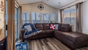 A cozy living room with a brown leather sectional sofa, a ceiling fan, and a large window with blinds. - Island Getaway• Patio View• Boat Ramp & Slips • Pets (Lake Havasu City)
