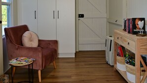 Ein Zimmer mit Holzboden, einem braunen Sessel mit Kissen, einem kleinen Tisch mit Zeitschriften, einem weißen Kleiderschrank mit schwarzen Griffen und einem Holzregal mit Büchern und Körben.