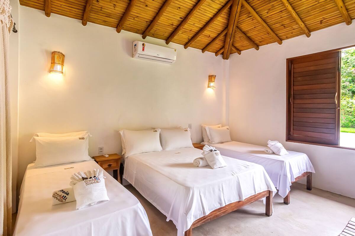 Quarto de hotel com duas camas, roupa de cama branca, teto de madeira e janela com persianas de madeira.