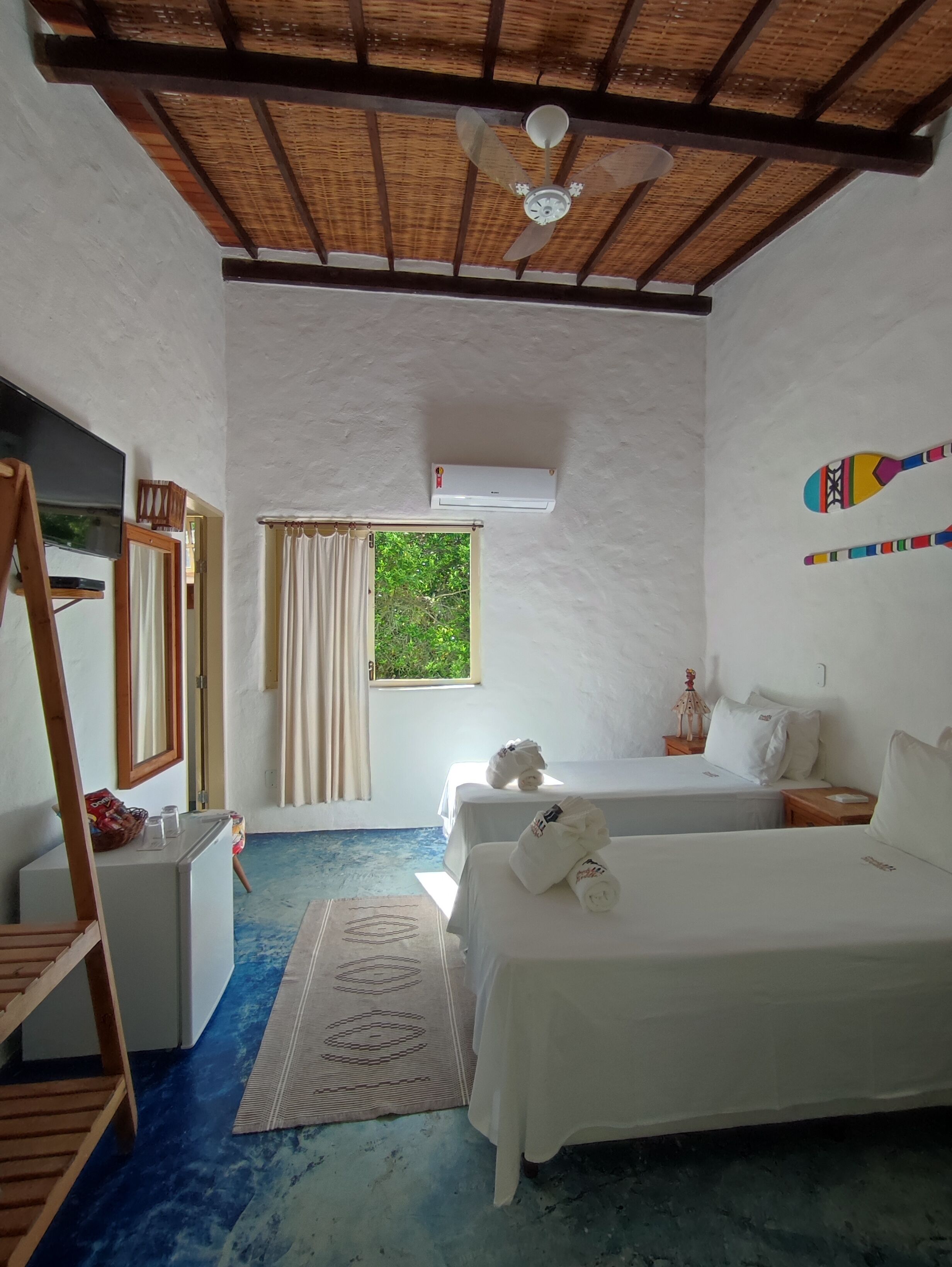 Une chambre avec deux lits, un ventilateur de plafond, un appareil de climatisation, une télévision et une fenêtre avec des rideaux.