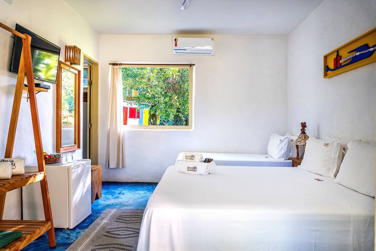 Quarto com cama, escrivaninha com computador, janela com vista para o verde e ar condicionado fixado na parede.