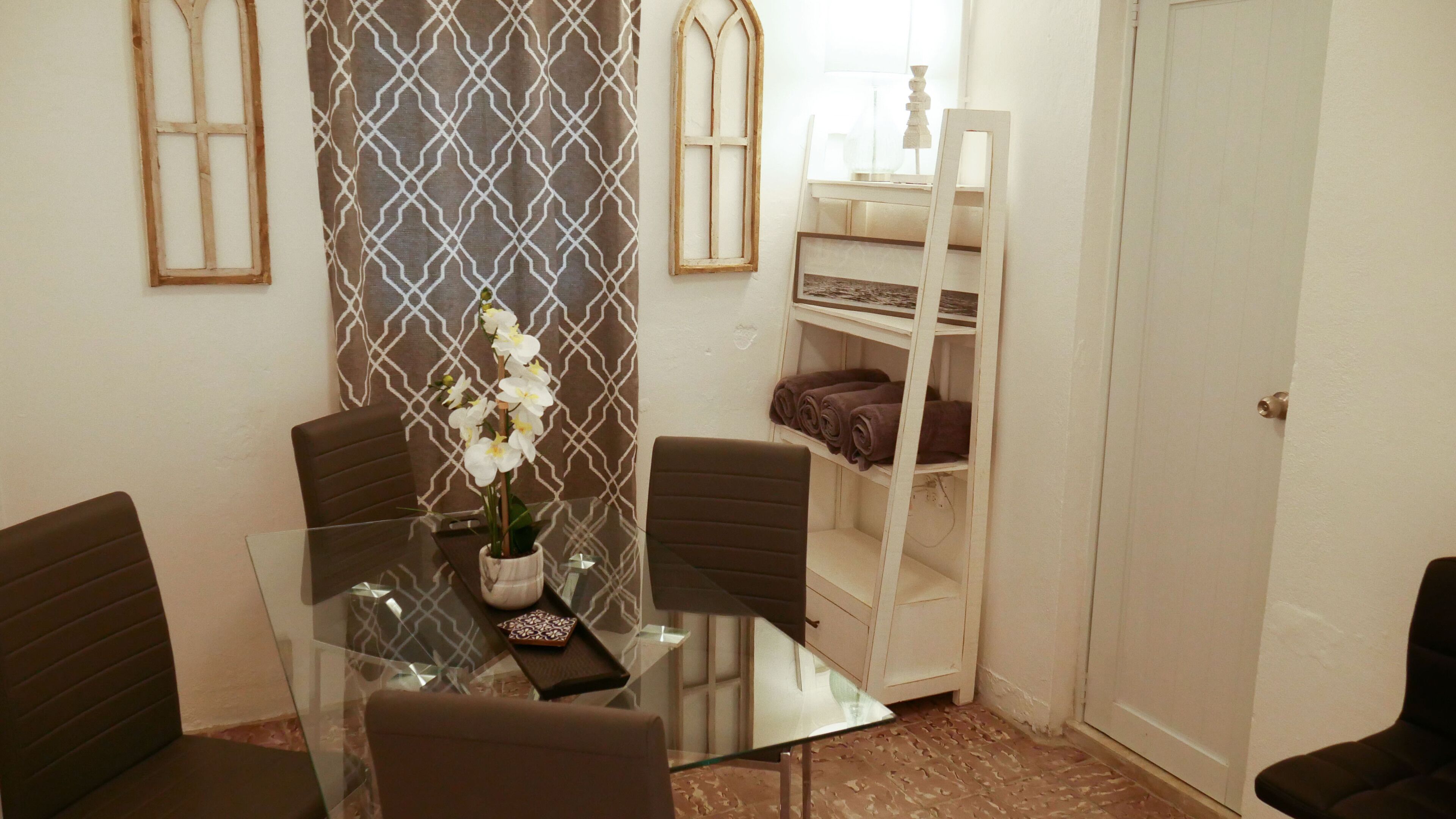 Una habitación con una mesa de vidrio, dos sillas, una orquídea en maceta, una cortina estampada, un estante blanco con toallas y una puerta.