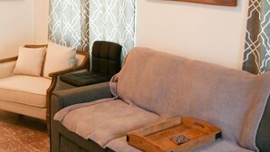 Een woonkamer met een grijze bank, een kleinere fauteuil, een houten dienblad en een plafondventilator.