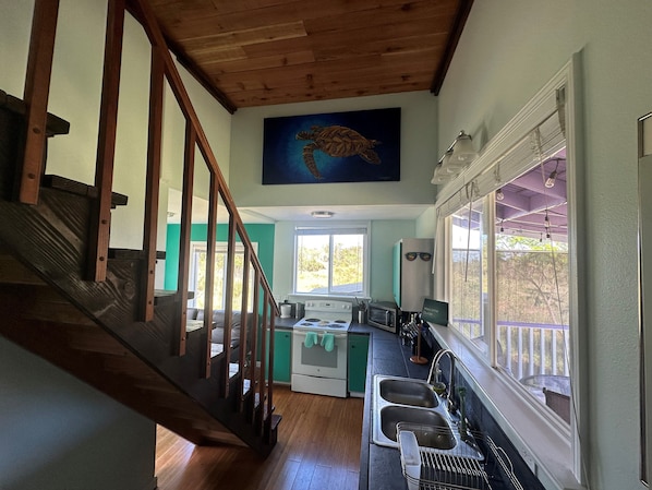 Una cocina con una escalera de madera, un cuadro sobre el fregadero y vistas al exterior a través de una ventana.