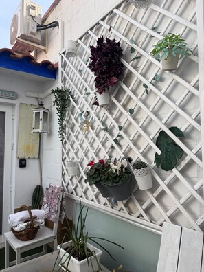Pared blanca con celosía, varias plantas en macetas, una cesta de mimbre con toallas y una pequeña mesa.