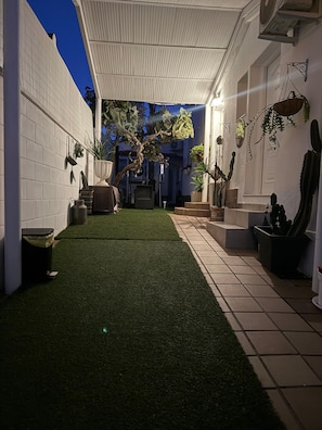 En overdækket terrasse med kunstgræs, plantekasser og udsigt til et hus.