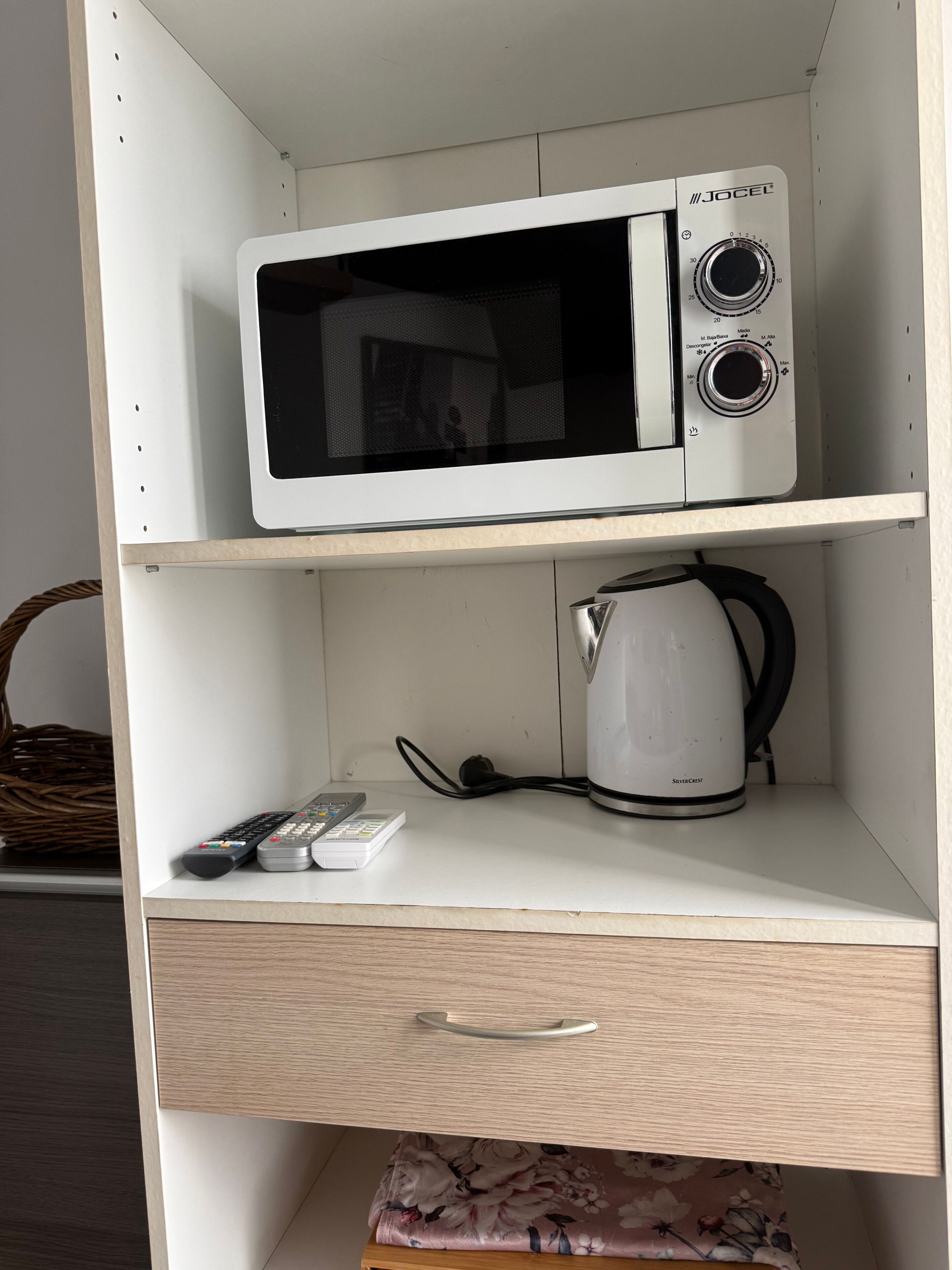A white microwave on a shelf, a kettle, a remote control, and a drawer with a floral pattern.