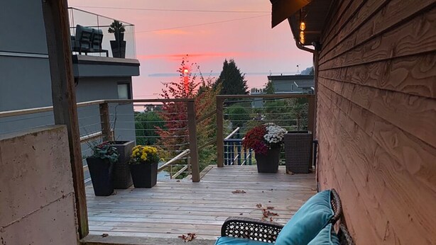 Un balcon avec un canapé en osier et une table en verre, donnant sur une terrasse en bois agrémentée de plantes en pot et offrant une vue sur le coucher de soleil.