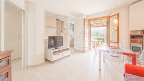 A modern living room with a TV, a glass dining table, and a kitchen area with a red oven. - Modern Casa Lula with double bathroom, garden and terrace immersed in the greenery of Marina di Camp (Campo nell'Elba)