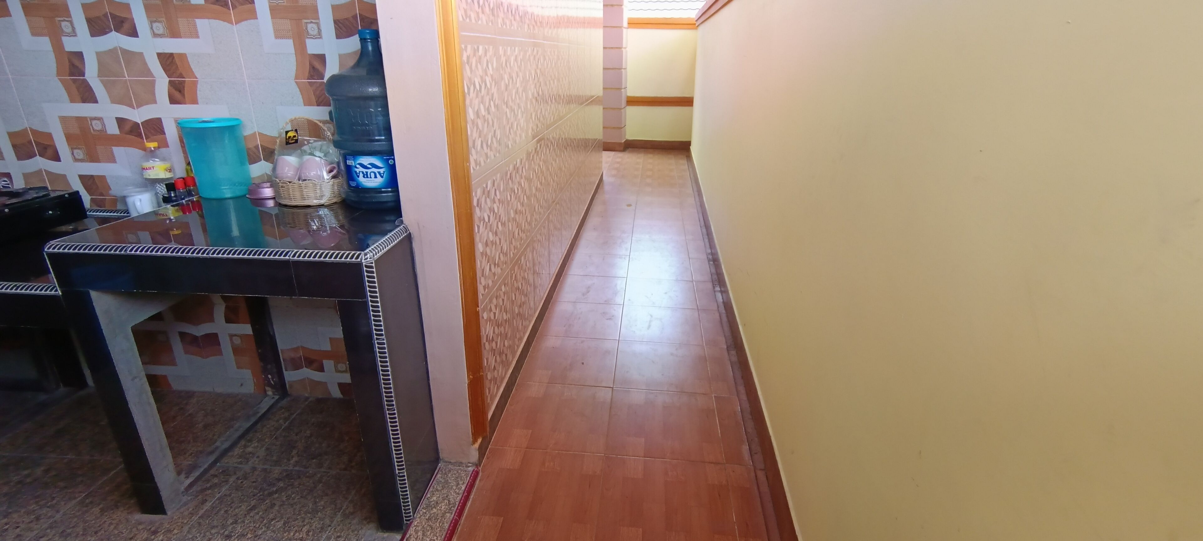 A narrow hallway with a tiled floor, a table with a water dispenser and other items, and a patterned wall.
