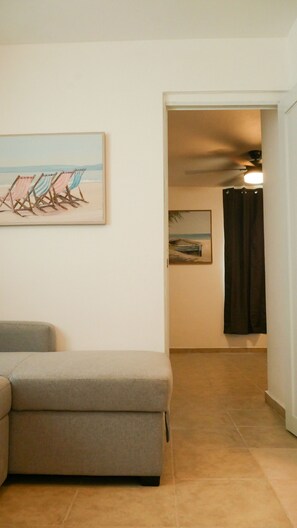 A living room with a grey sofa, a painting of beach chairs, and a hallway leading to a bedroom with a bed and a ceiling fan.