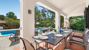 Un área de comedor al aire libre con vista a la piscina, mobiliario exterior y una pérgola.