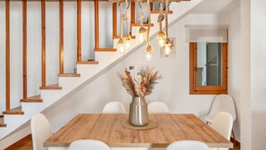 Un comedor con una mesa de madera, sillas blancas, un jarrón con flores secas y una escalera con barandillas de madera y luces colgantes.