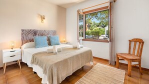 Un dormitorio con cama, mesitas de noche, una silla de madera, una ventana con vista a follaje y una lámpara fijada a la pared.