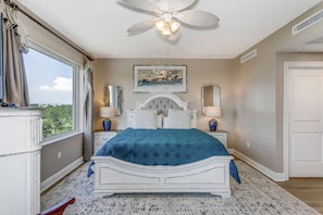 Une chambre avec un grand lit, un ventilateur de plafond, une fenêtre avec des rideaux, un tableau au mur et une porte.