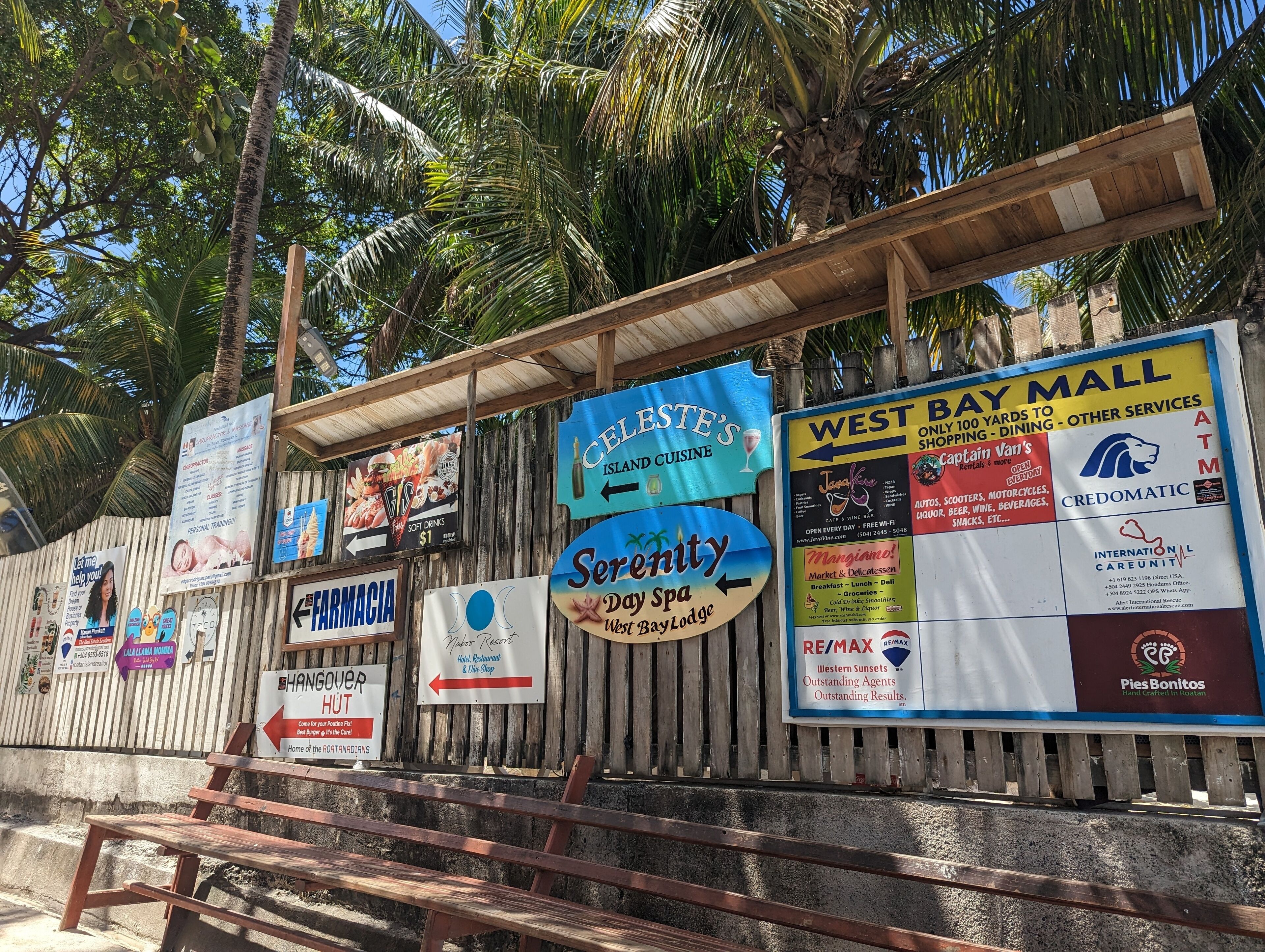 Une clôture en bois ornée de divers panneaux publicitaires pour des commerces proposant des services tels qu’une pharmacie, un spa et un centre commercial.