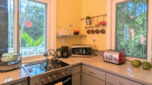 Une cuisine compacte équipée d’une cuisinière à gaz, d’un four à micro-ondes, d’un grille-pain et d’une machine à café. Deux fenêtres laissent entrer la lumière naturelle et offrent une vue sur la verdure environnante.