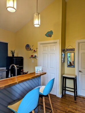 A compact kitchen with a wooden bar, two blue chairs, a refrigerator, and a door with a sign.