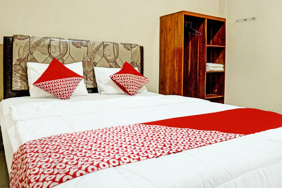 A hotel room with a large bed, two pillows with a red and white pattern, a red throw blanket, and a wooden wardrobe with shelves.