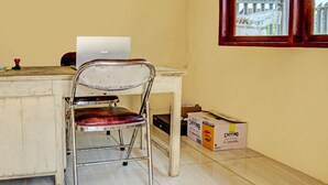 Een kleine kamer met een houten bureau, een metalen stoel, een laptop, een doos CaffinB en een raam met uitzicht op een hek.
