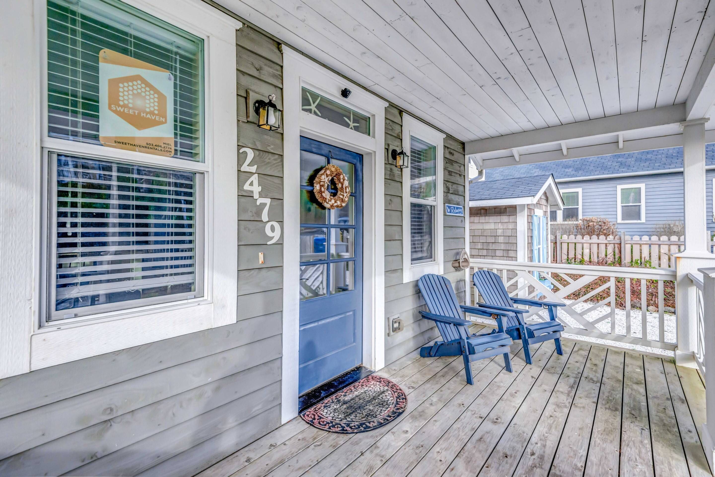 A wooden porch with a blue door, two blue Adirondack chairs, and a decorative wreath.