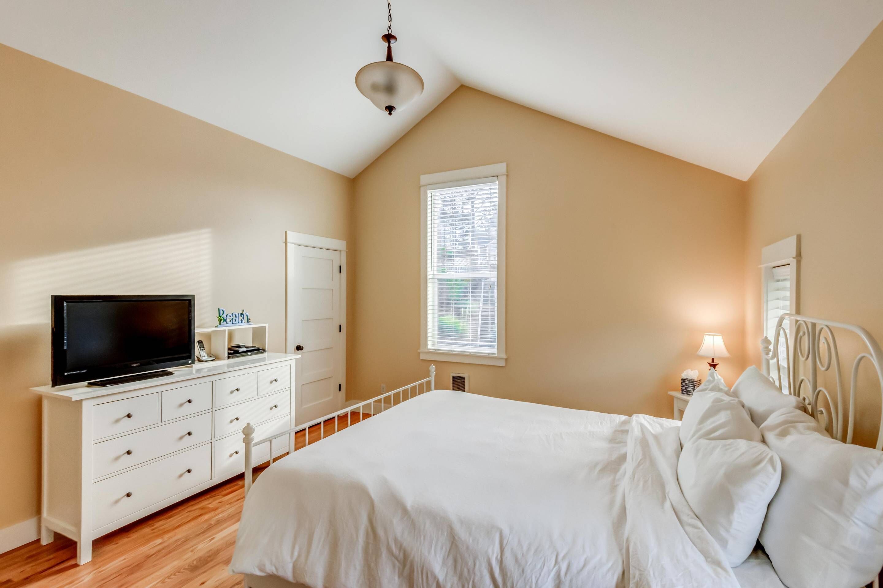 A bedroom with a bed, a TV on a dresser, a window with blinds, and a bedside lamp.
