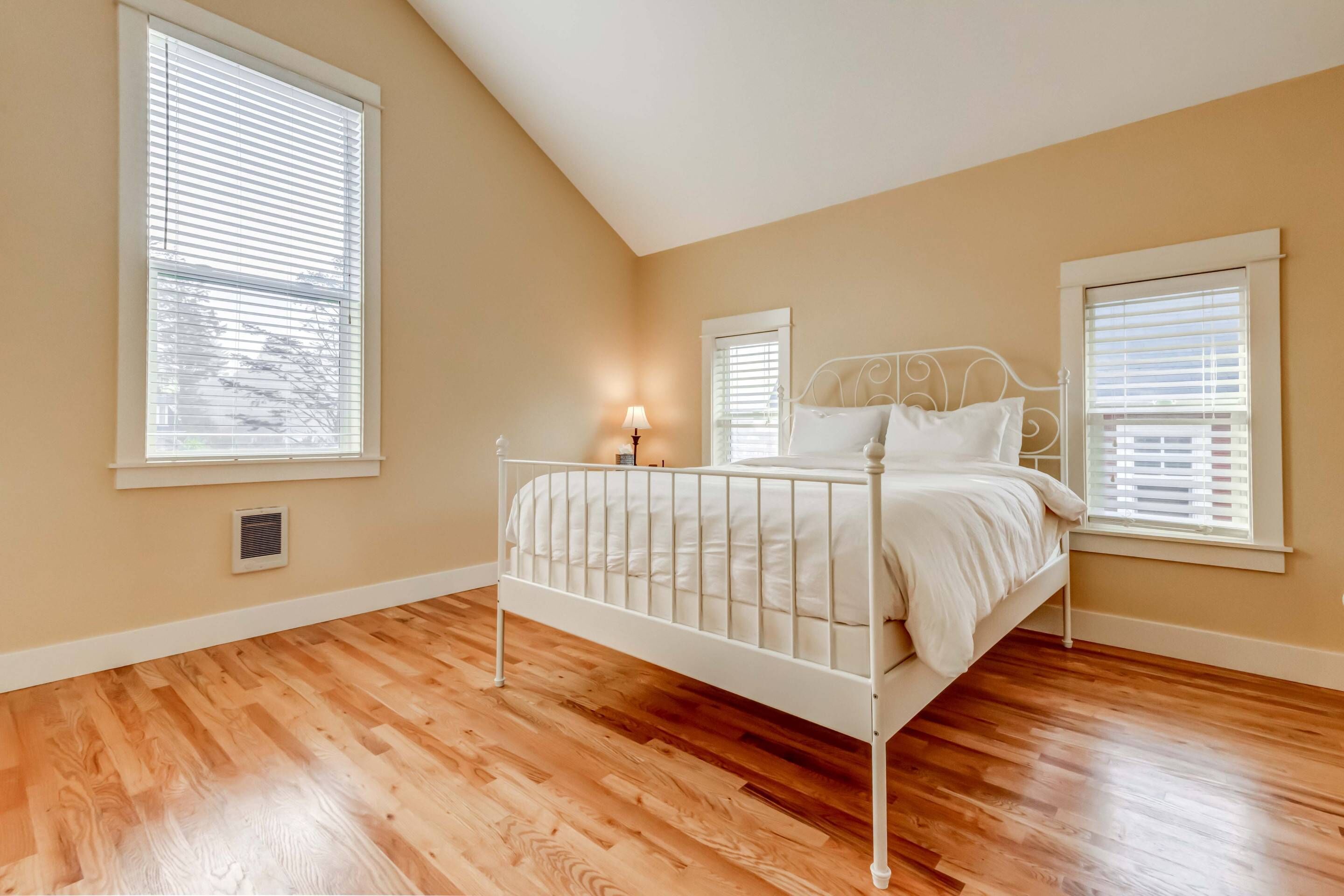A bedroom with a white metal bed frame, wooden flooring, and two windows with blinds.