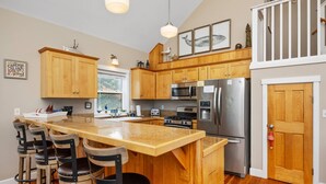 A kitchen with wooden cabinets, a stainless steel refrigerator, a microwave, and a stove. There are three stools at the island and a window above the sink.