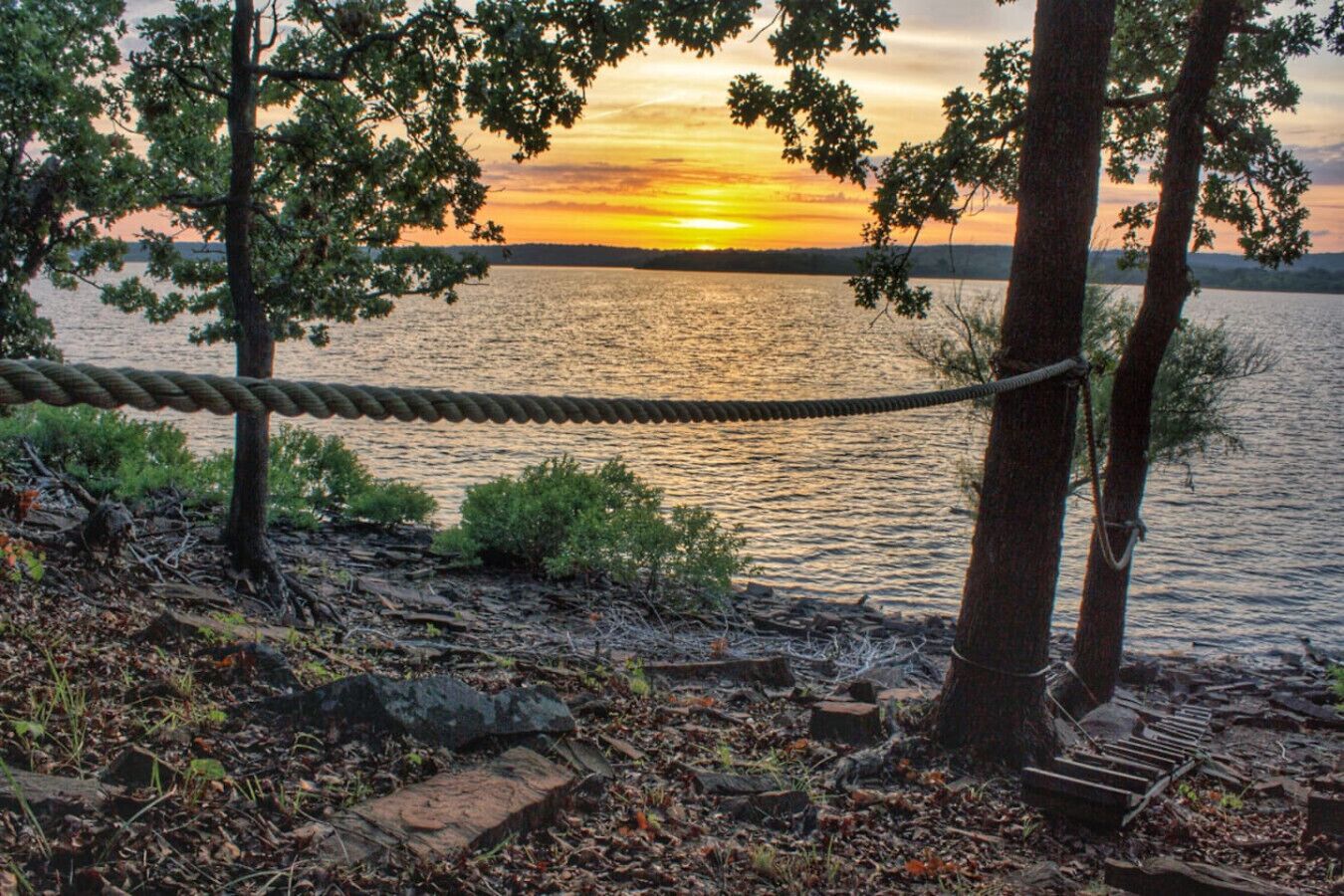Köysiliuku kivikkoisen rantaviivan yllä, taustalla puita ja auringonlasku veden päällä.