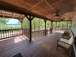 Eine überdachte Holzterrasse mit Blick auf einen See und Bäume, Holzstühle und ein Deckenventilator.