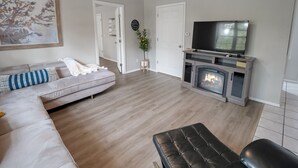 Un salon avec un canapé gris, un pouf noir, une cheminée, un téléviseur et un ventilateur de plafond.