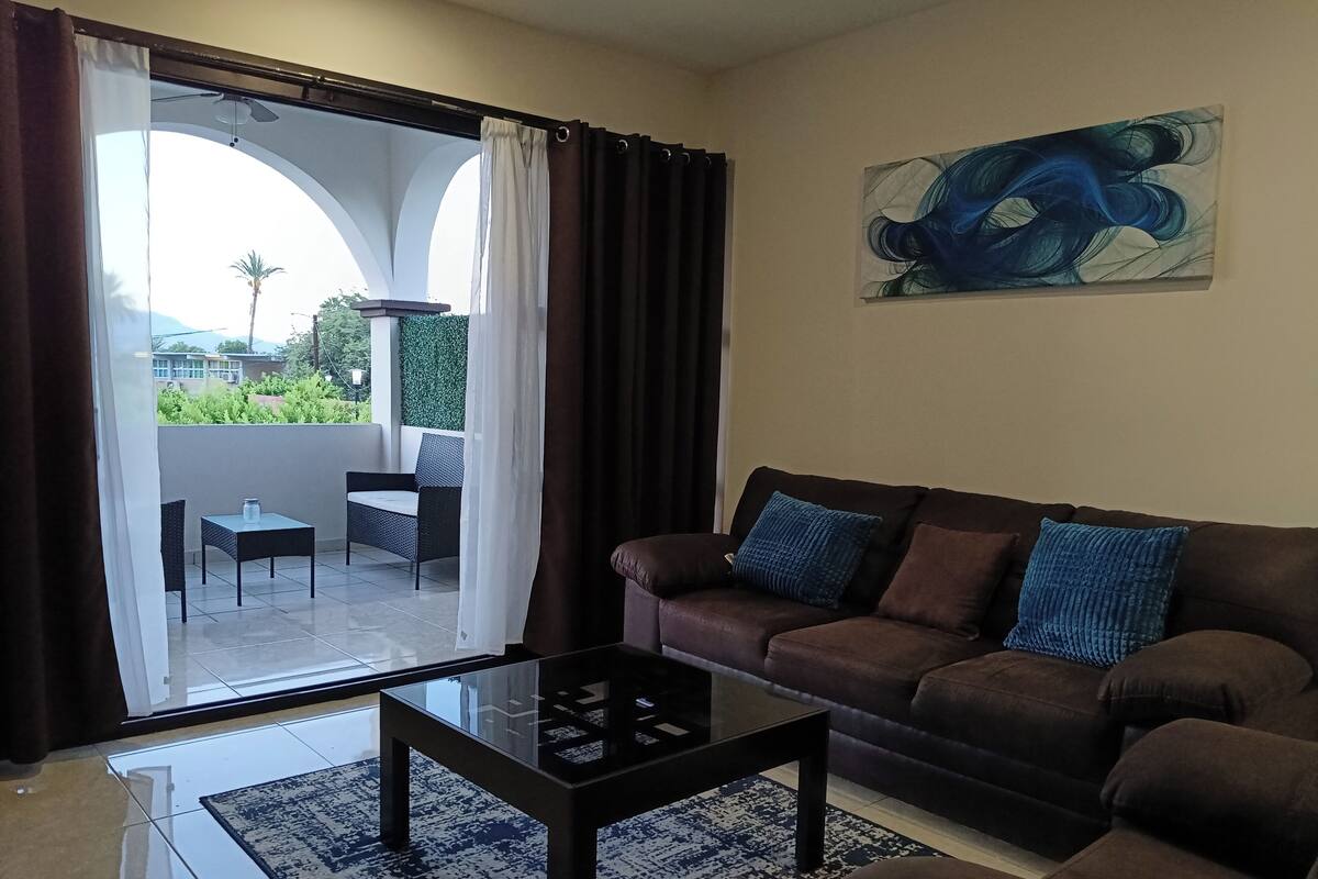Una sala de estar con un sofá marrón, una mesa de centro de vidrio y vistas a un balcón con mesa y sillas.