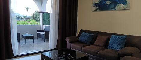 Una sala de estar con un sofá marrón, una mesa de centro de vidrio y vistas a un balcón con mesa y sillas.