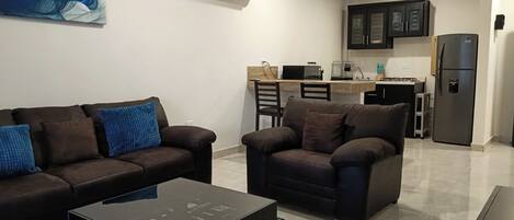 Una sala de estar moderna con sofá, sillón y mesa de centro, un área de cocina con armarios y electrodomésticos, y un cuadro en la pared.