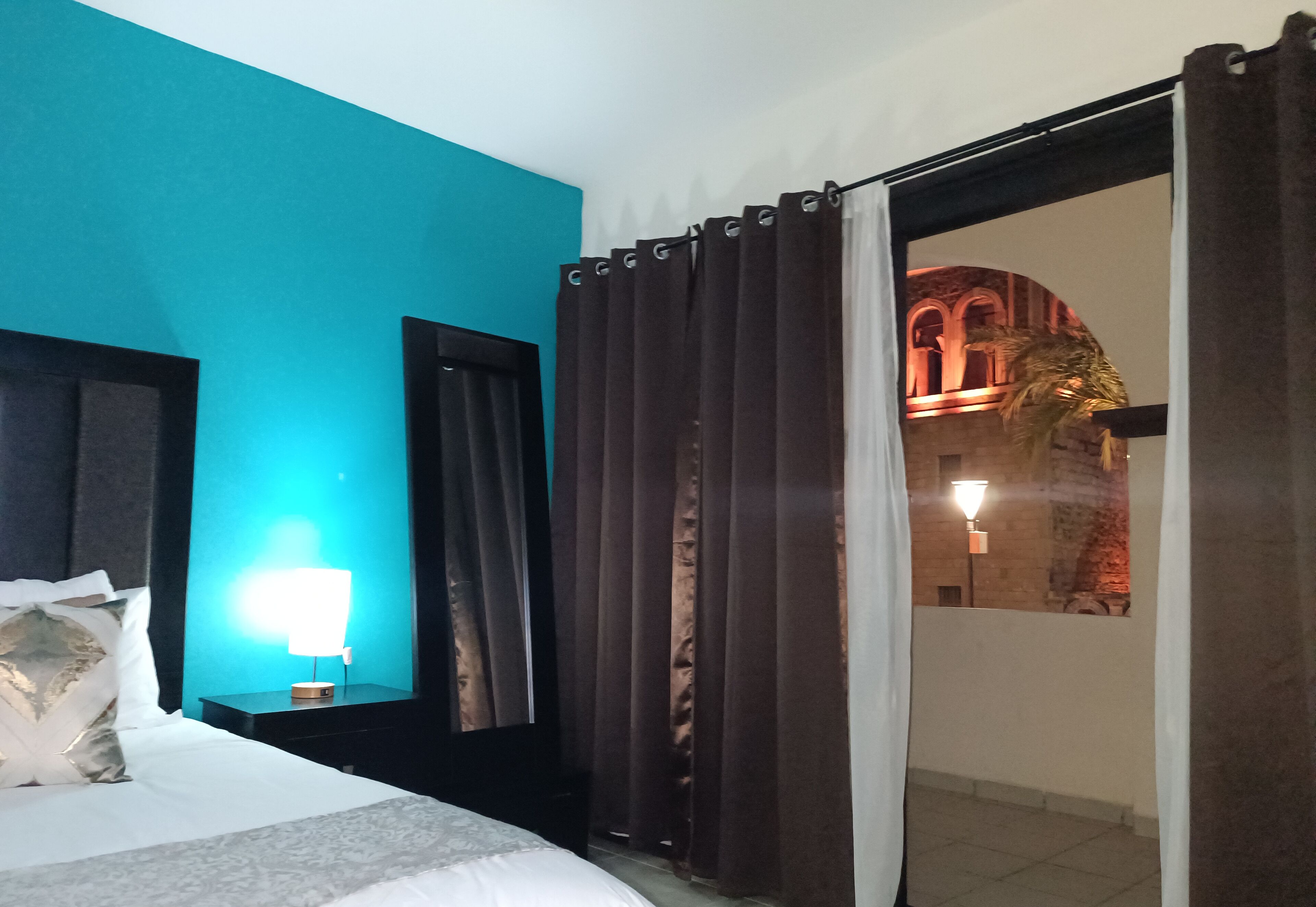 Habitación de hotel con cama, mesita de noche con lámpara, ventana con cortinas y vista a un edificio.