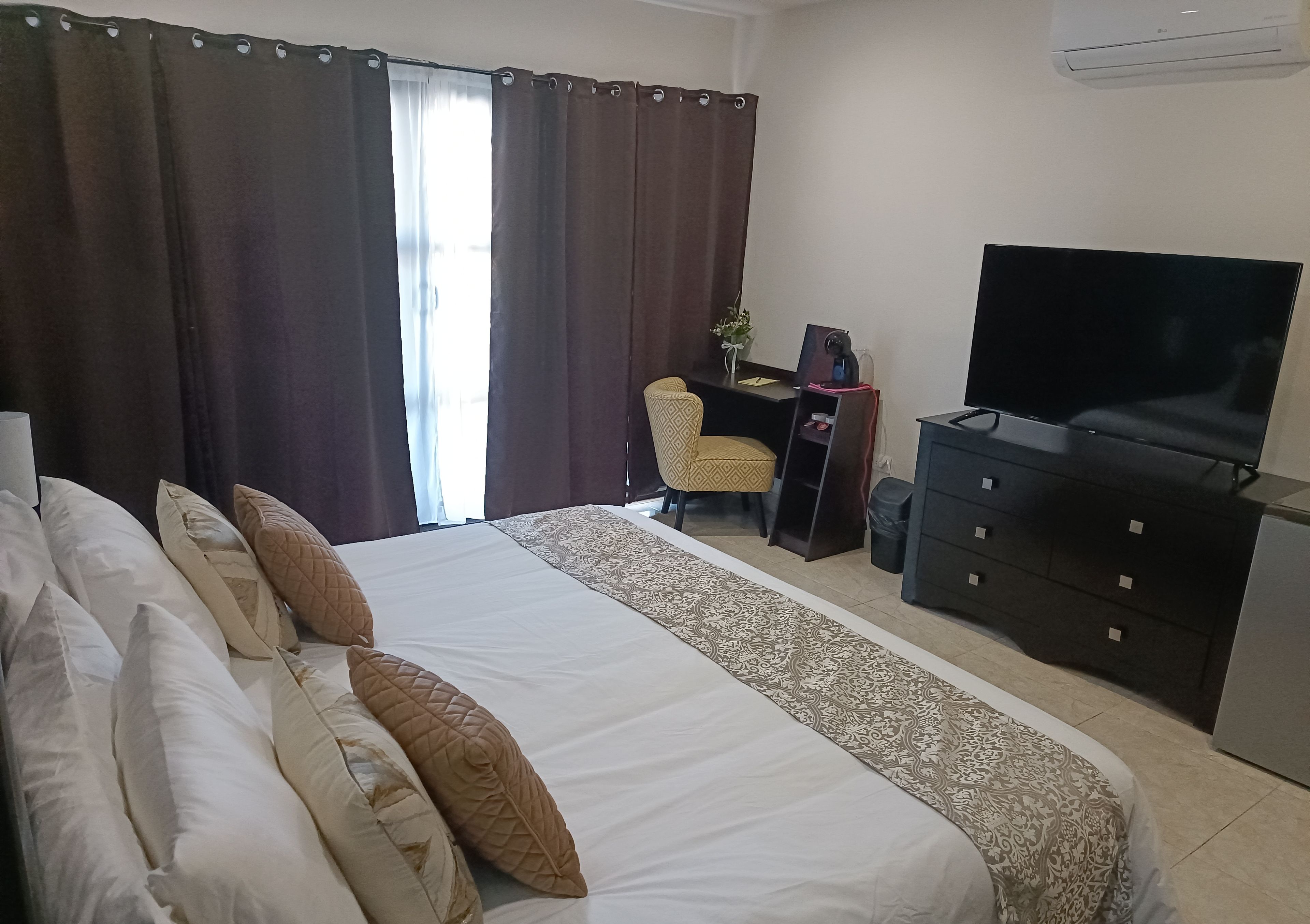 A hotel room with a large bed, a desk with a chair, a television, and an air conditioning unit.