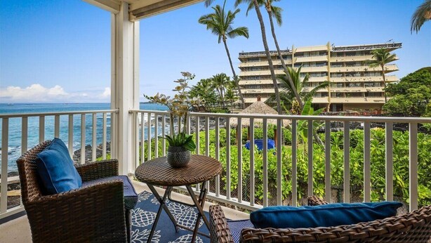 A balcony with wicker furniture, a small table, and a view of the ocean and a building with palm trees.