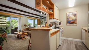 A compact kitchen with a central island, a dining area with a sofa and table, and a large window offering a view of greenery.