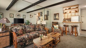 A living room with a sofa, chairs, a coffee table, a TV, and a kitchen area with a fridge and bar stools.