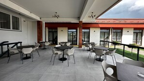 An outdoor patio area with multiple tables and chairs, large windows, and a red-roofed building in the background.