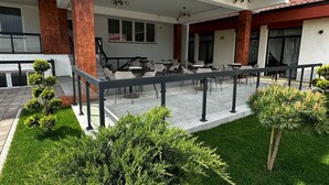 Un espacio exterior moderno con mesas y sillas, rodeado de césped cuidado y pequeños árboles.