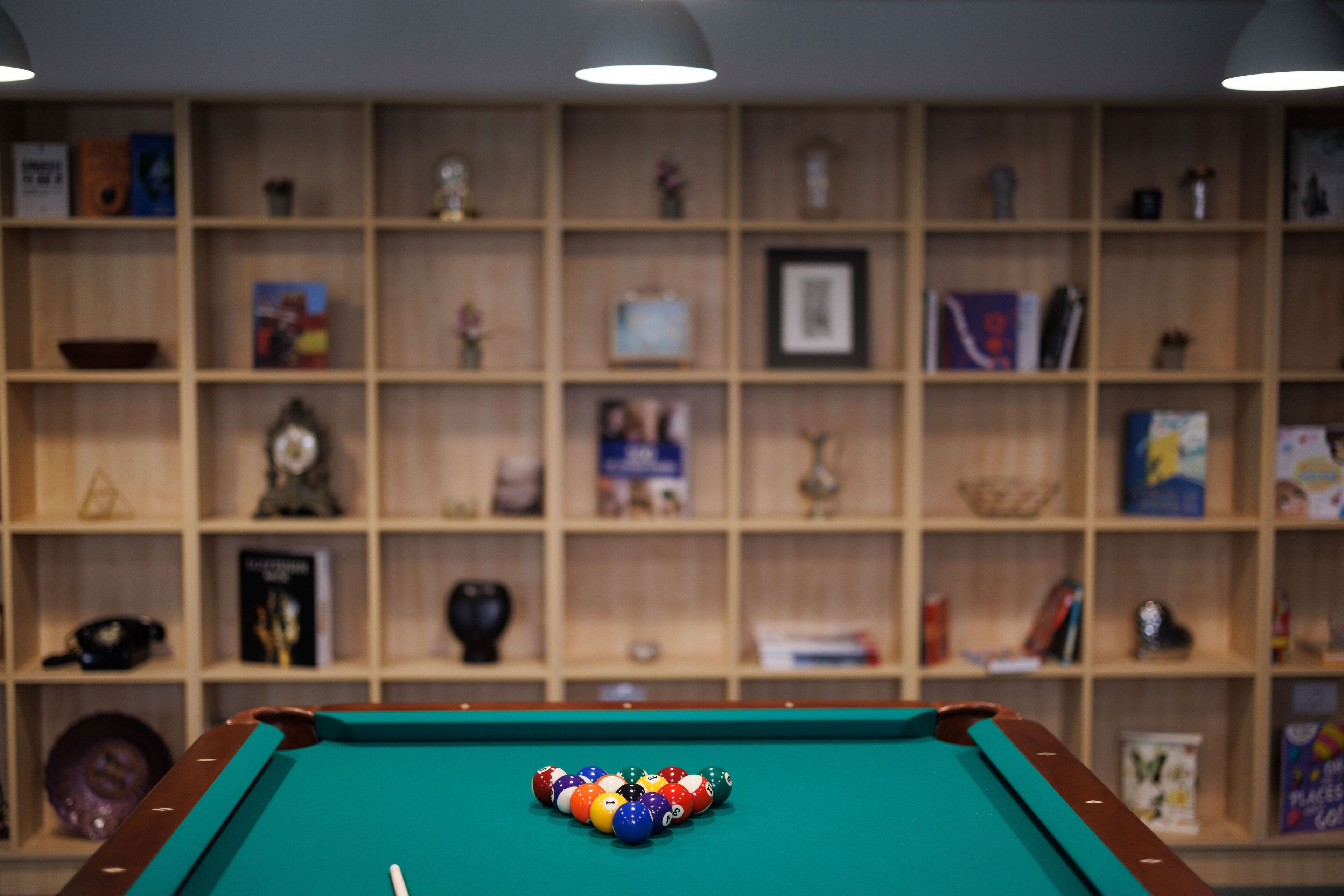 Una mesa de billar con bolas en el centro, rodeada por una estantería de madera llena de objetos varios.