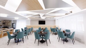 A modern dining area with geometric ceiling design, multiple tables and chairs, and a kitchen in the background.