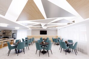 A modern dining area with geometric ceiling design, multiple tables and chairs, and a kitchen in the background.