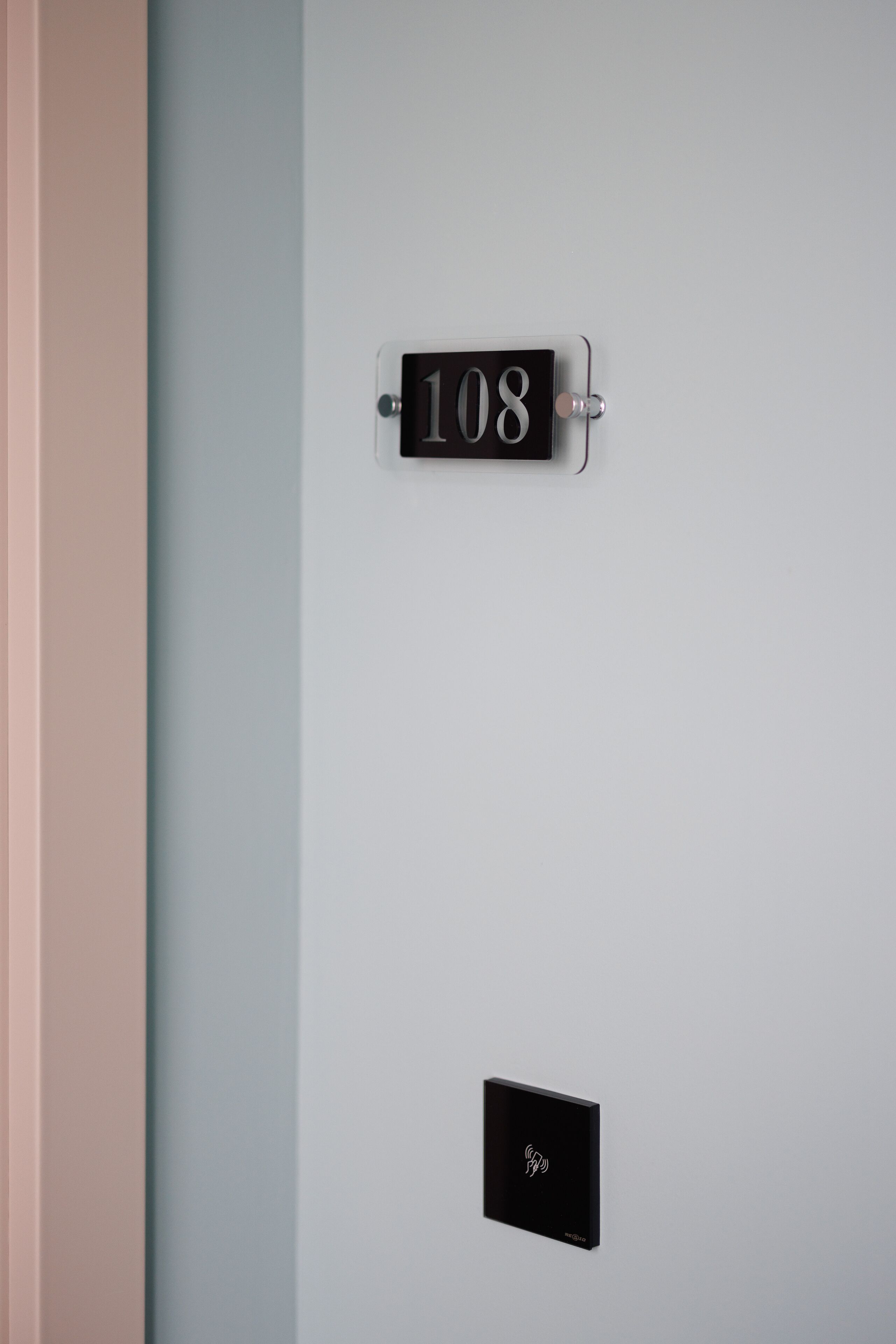 Una placa con el número 108 montada en la pared, un pequeño panel de control debajo y un marco de puerta a la izquierda.